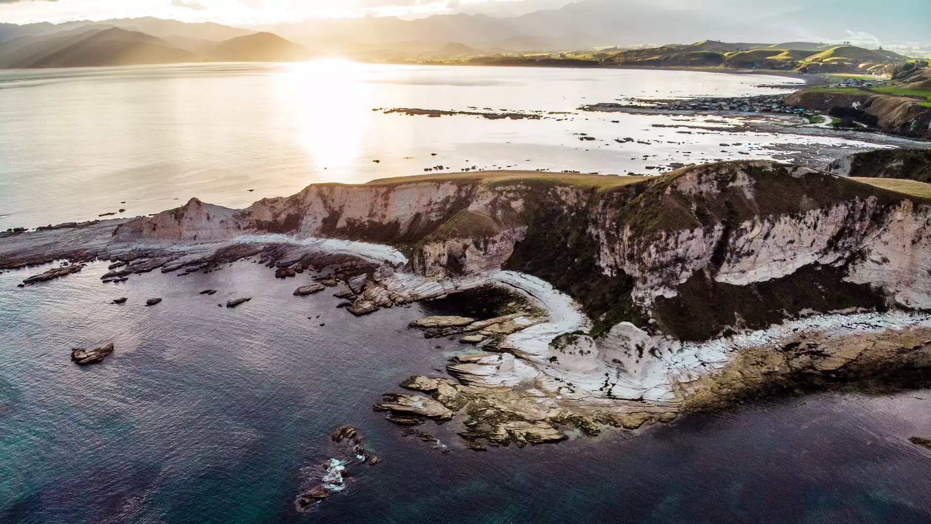 A high-altitude drone view of white limestone cliffs and a rocky reef jutting into a dark teal ocean, with three tiny yellow kayaks in the water and a bright sun glare on the horizon.