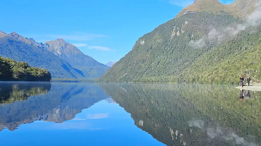 Calm lake reflecting green mountains and blue sky with two people standing by the shore