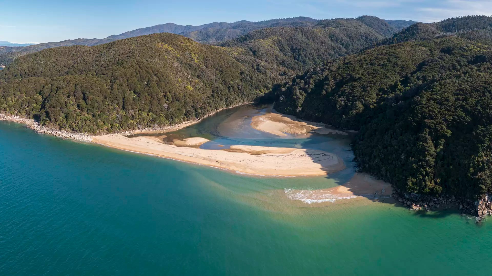 A high-altitude aerial view of a river winding through golden sand to meet a turquoise sea, bordered by lush green forest and a rugged coastline under a clear sky.