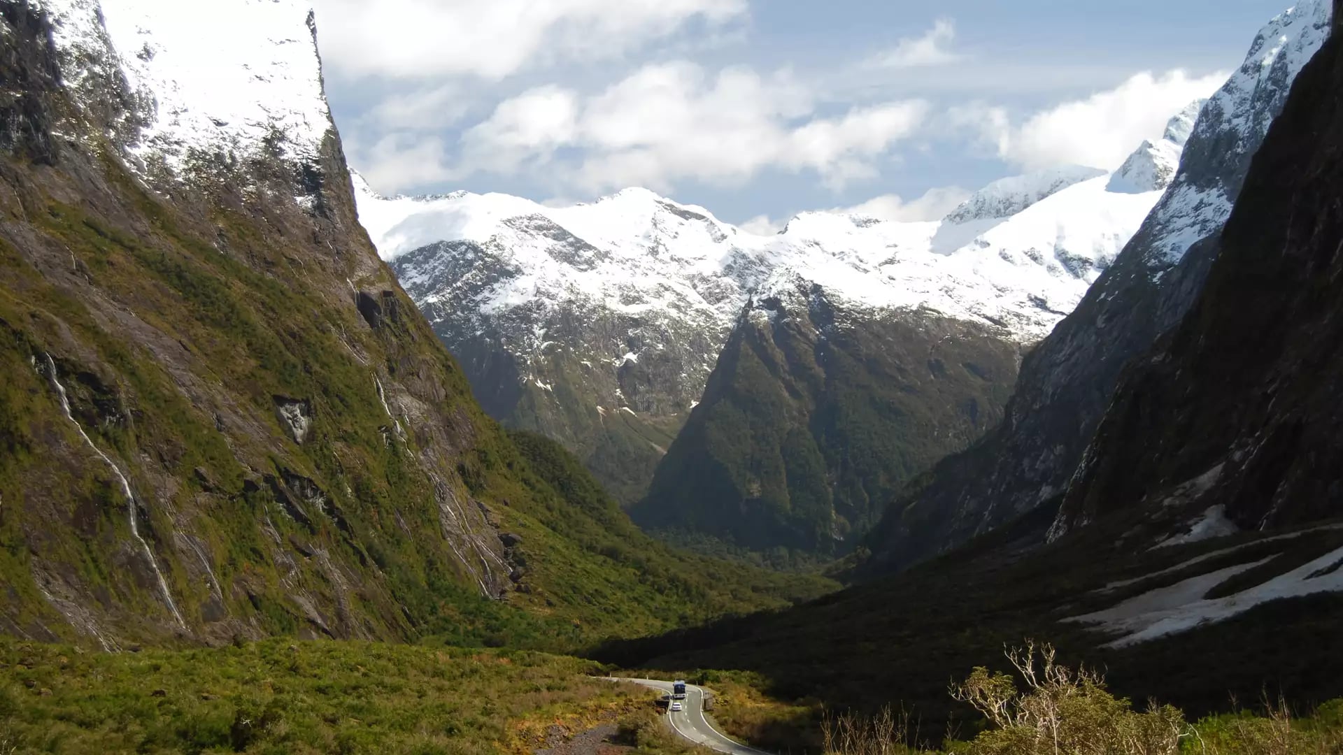 A winding highway snakes through the deep, green Cleddau Valley, surrounded by towering, snow-capped granite peaks and steep mountain walls in Fiordland.