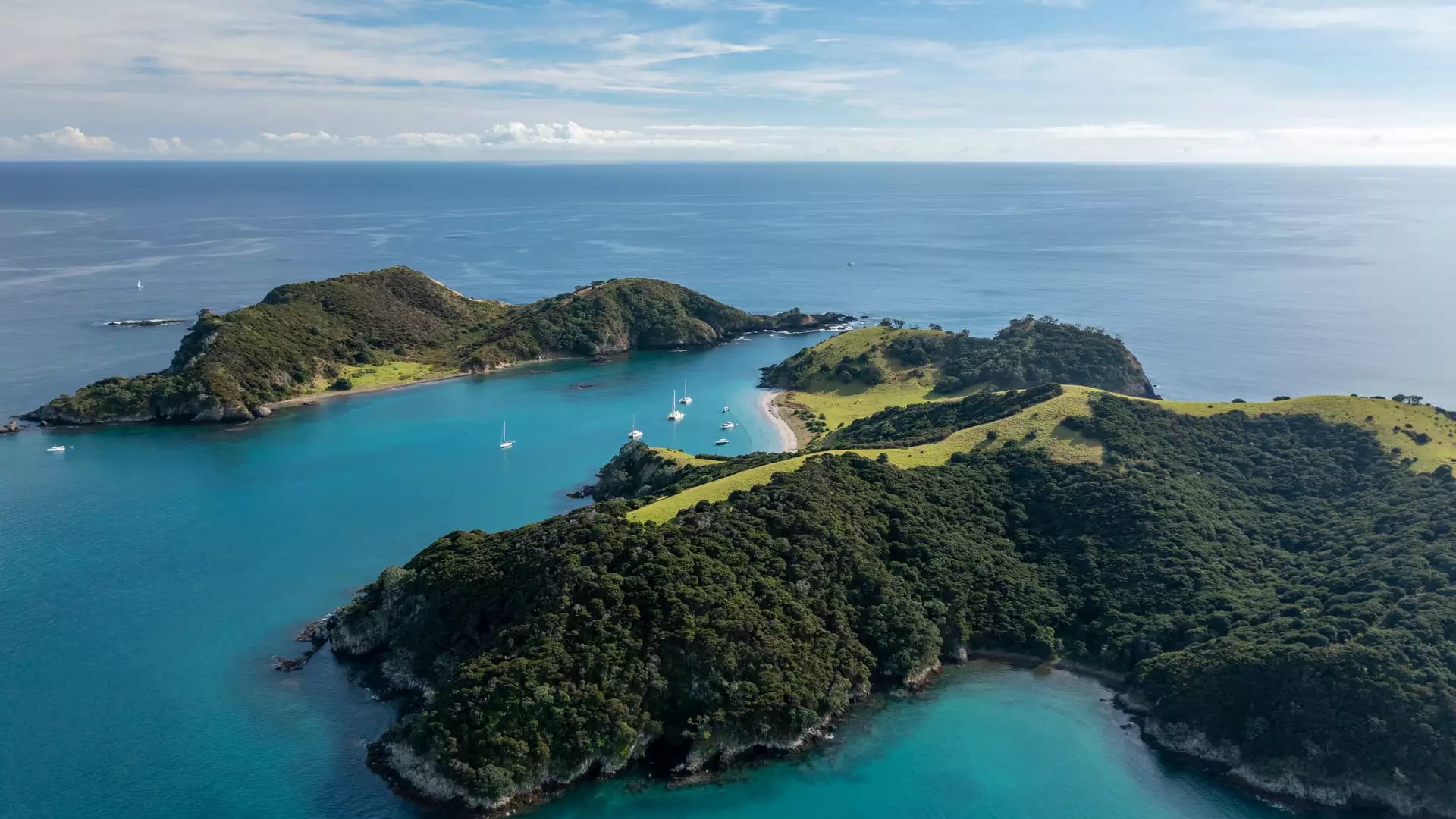 A high-altitude aerial view of Roberton Island in the Bay of Islands, featuring two bright turquoise lagoons separated by a narrow green isthmus with anchored sailboats.