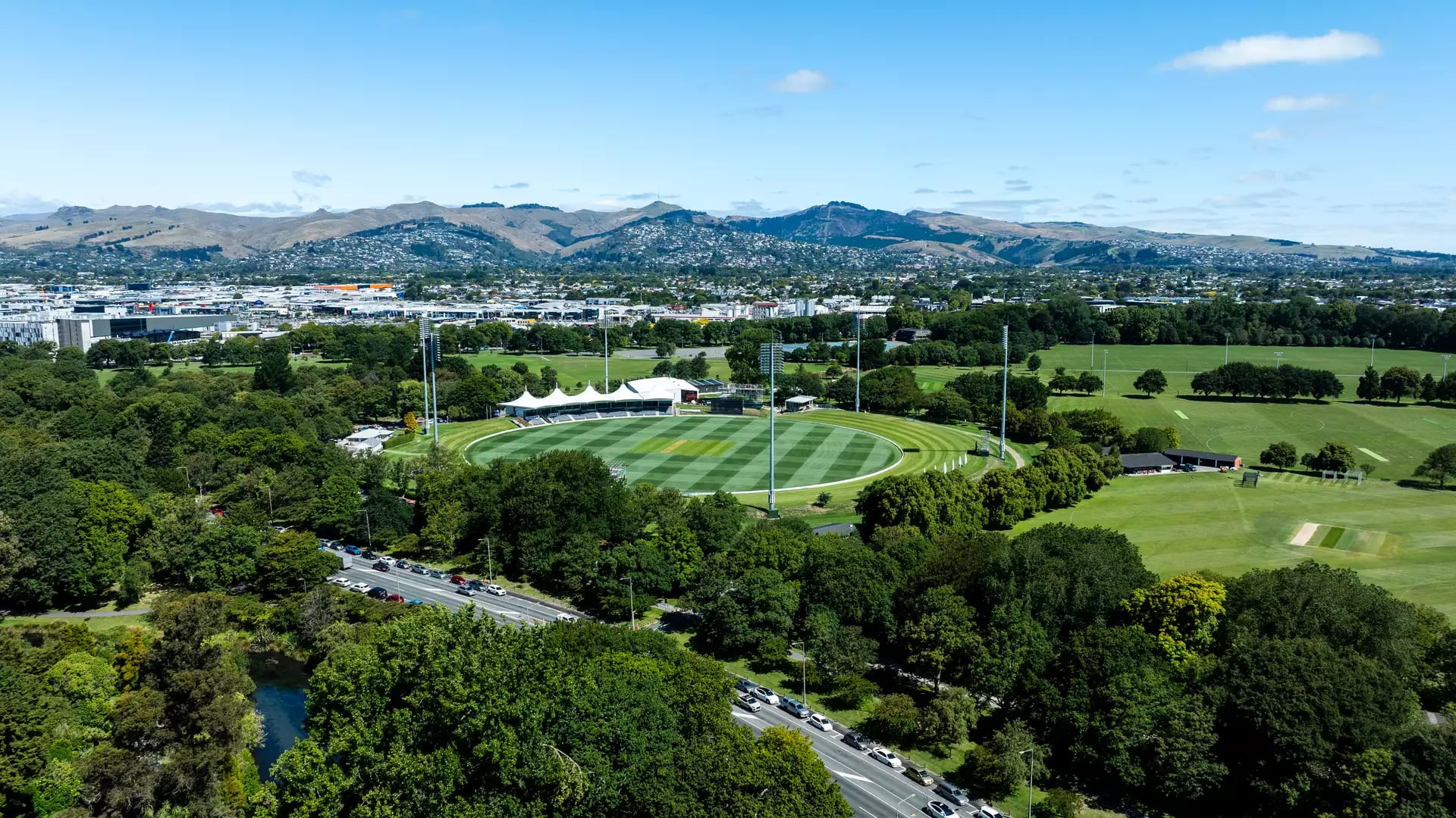 A high-altitude aerial view of a circular green cricket ground with a modern pavilion, surrounded by dense green forest and parkland, with a city and mountains in the background.