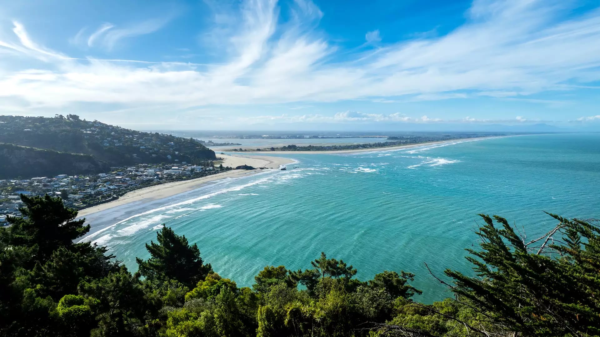 A high-altitude coastal view overlooking a turquoise bay and a long, straight sandy beach stretching into the distance, with a hillside suburb in the foreground under a sky with wispy white clouds.