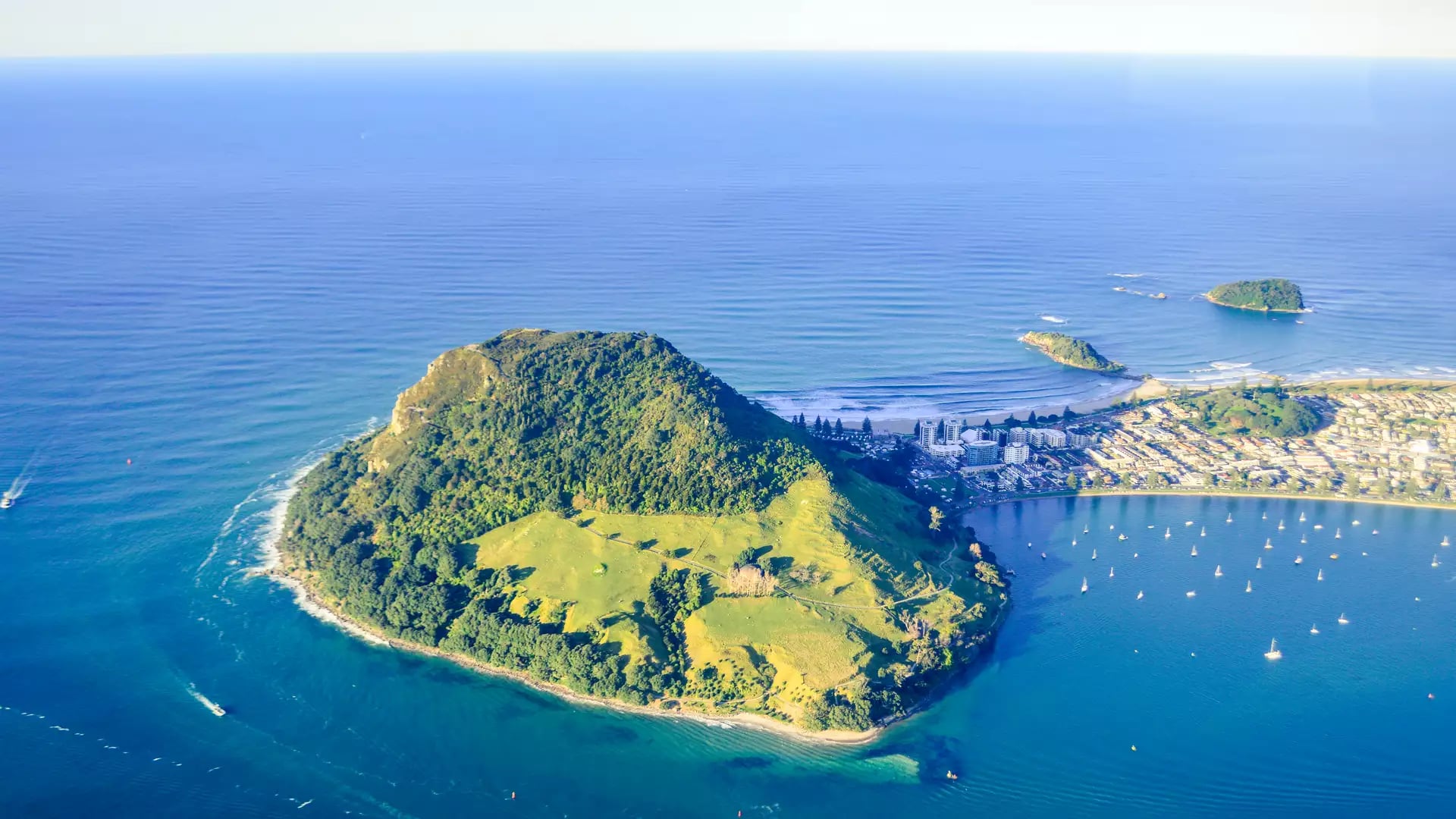 A high-altitude aerial shot of Mauao (Mount Maunganui) bathed in afternoon sunlight, showing the green volcanic cone, the white sand isthmus, and the turquoise waters of the Bay of Plenty filled with boats.