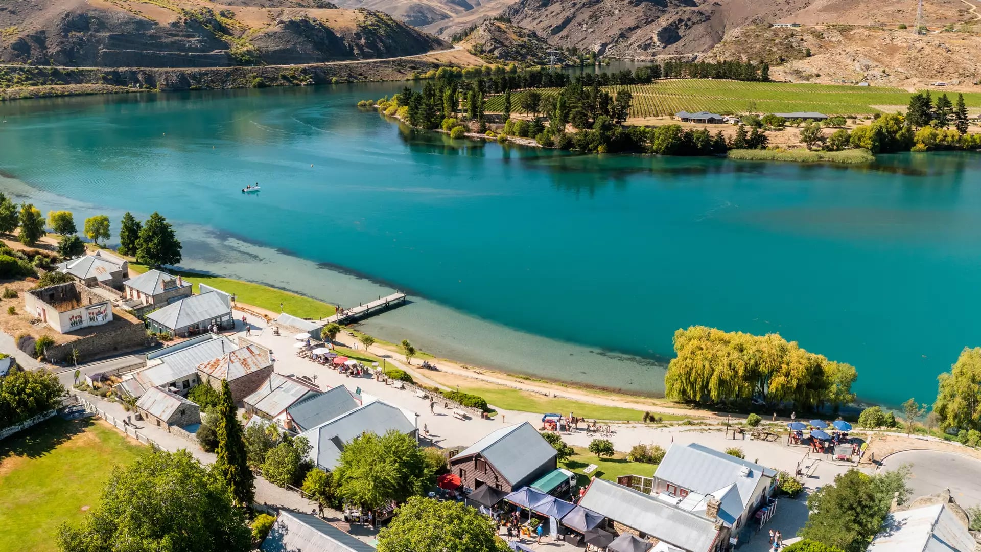A high-angle aerial view of a historic stone village with market stalls next to a vibrant turquoise lake, with vineyards and rugged dry mountains in the background.