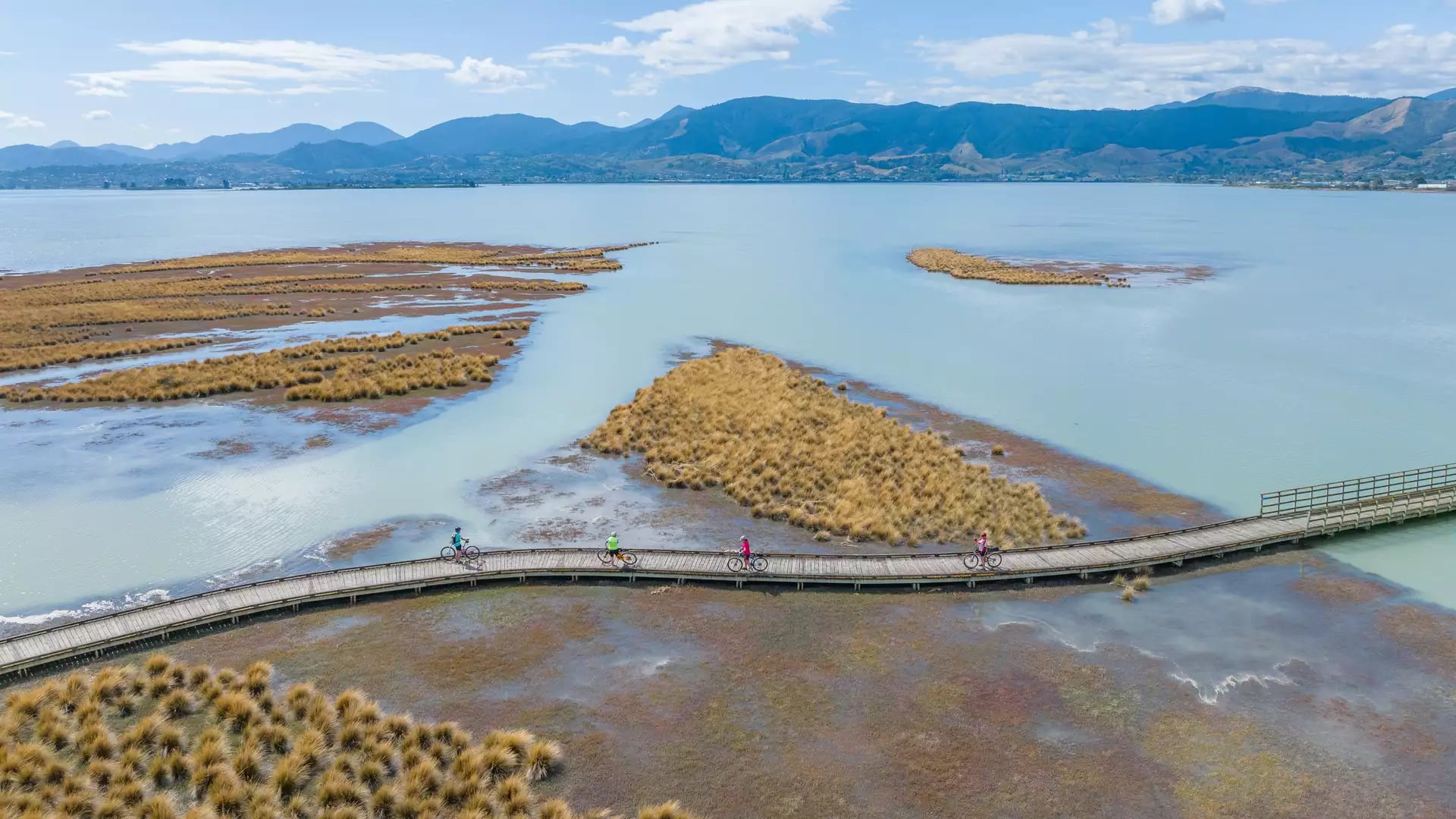 An aerial view of four cyclists riding across a long wooden boardwalk over a shallow coastal inlet with golden marsh grasses and distant blue mountains.