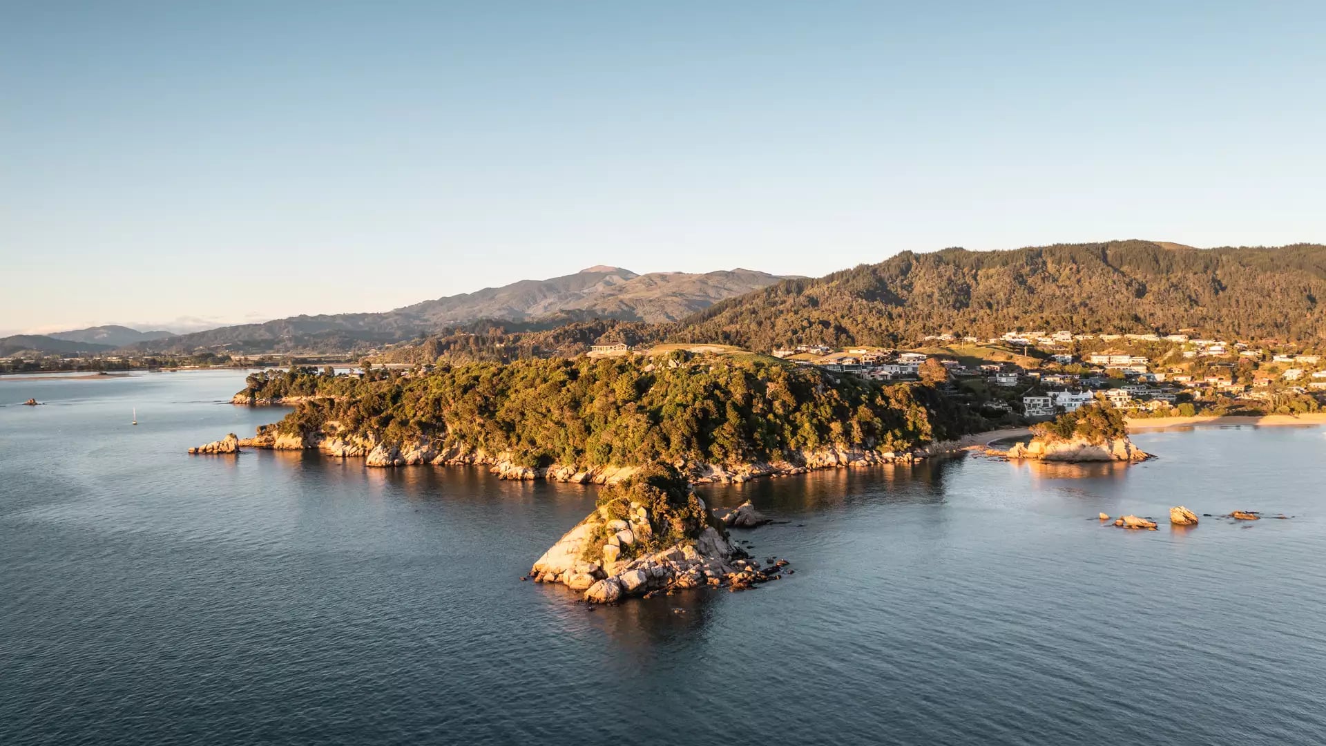 Aerial view of rocky coastline and beach with forested hills and houses under clear blue sky.
