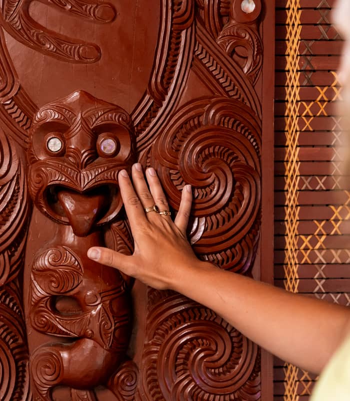 A person gently touches an intricate Māori wood carving (whakairo) featuring traditional spiral patterns and a central figure with paua shell eyes.