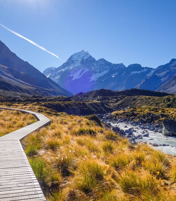 Boardwalk leading through grassland to snow-capped Aoraki Mount Cook under a bright blue sky.