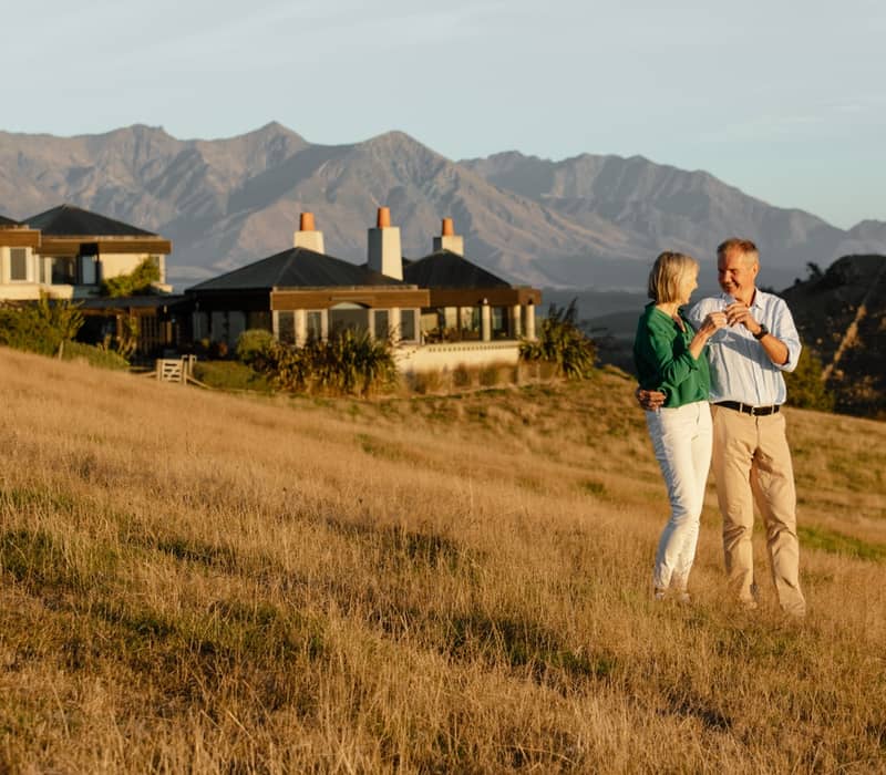 Couple standing on dry grass in front of a lodge with mountains at sunset.