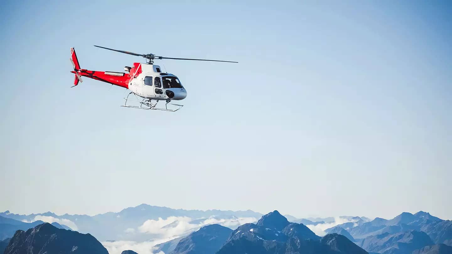 A red and white helicopter flies through a clear blue sky above a vast, jagged range of snow-capped mountains and white clouds.