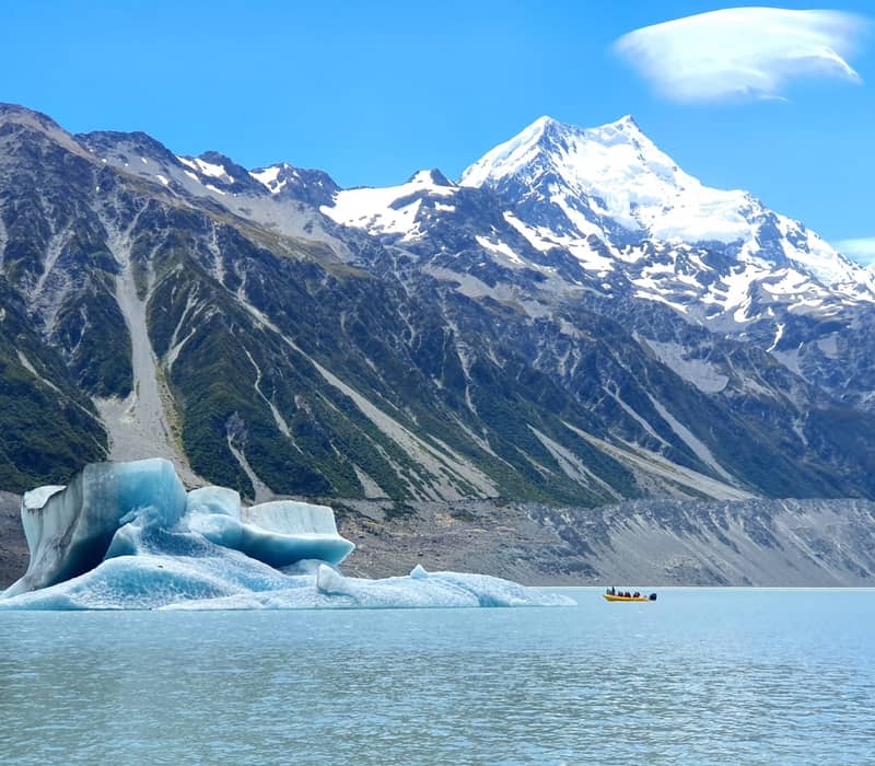 Boat with people near an iceberg on a lake with snow-covered Mount Cook in the background under a clear blue sky.