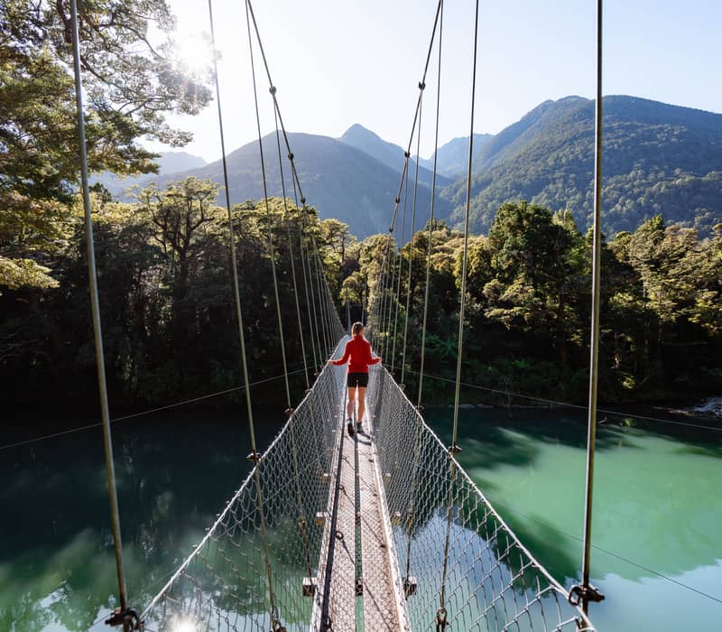 Person crossing suspension bridge over river in forested mountainous area under bright sun.