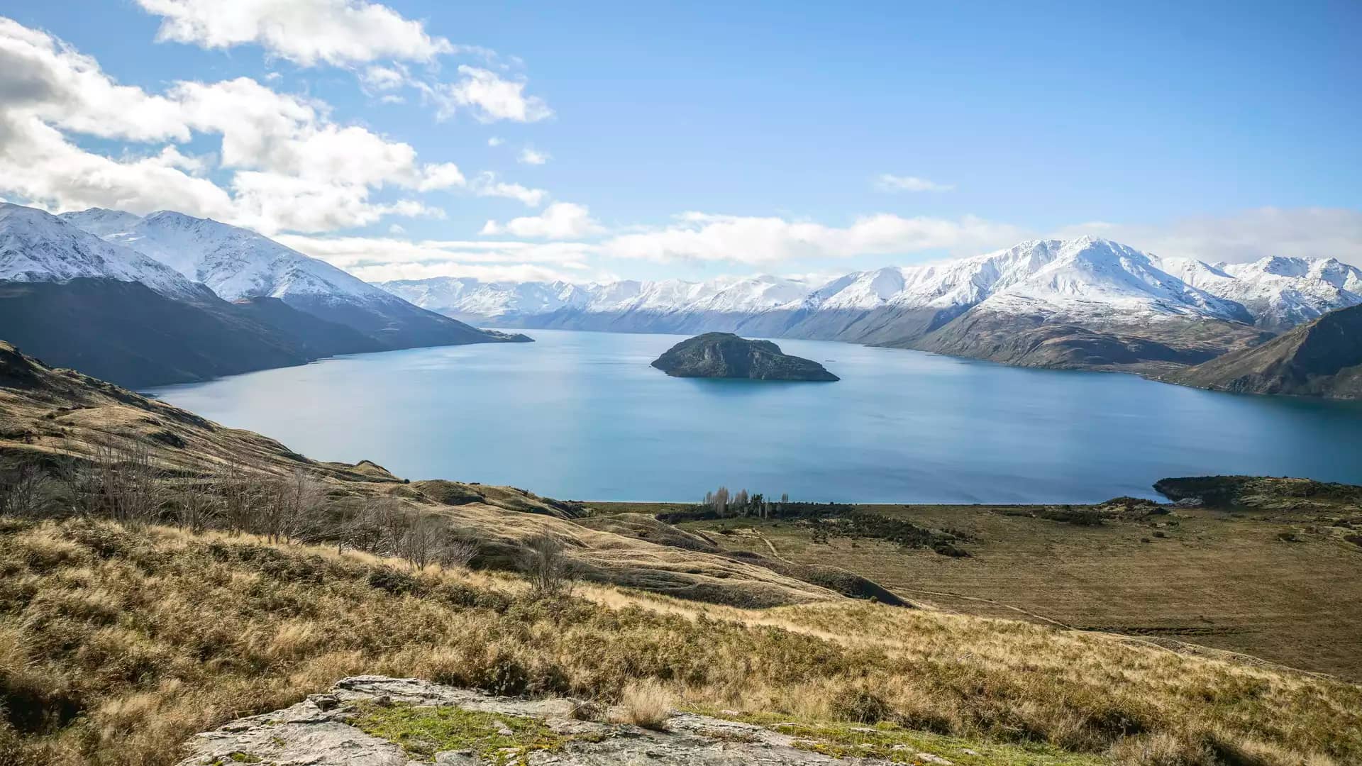 An expansive high-angle view of Lake Wānaka with a small green island in the center, surrounded by massive snow-covered mountain ranges under a bright blue sky.
