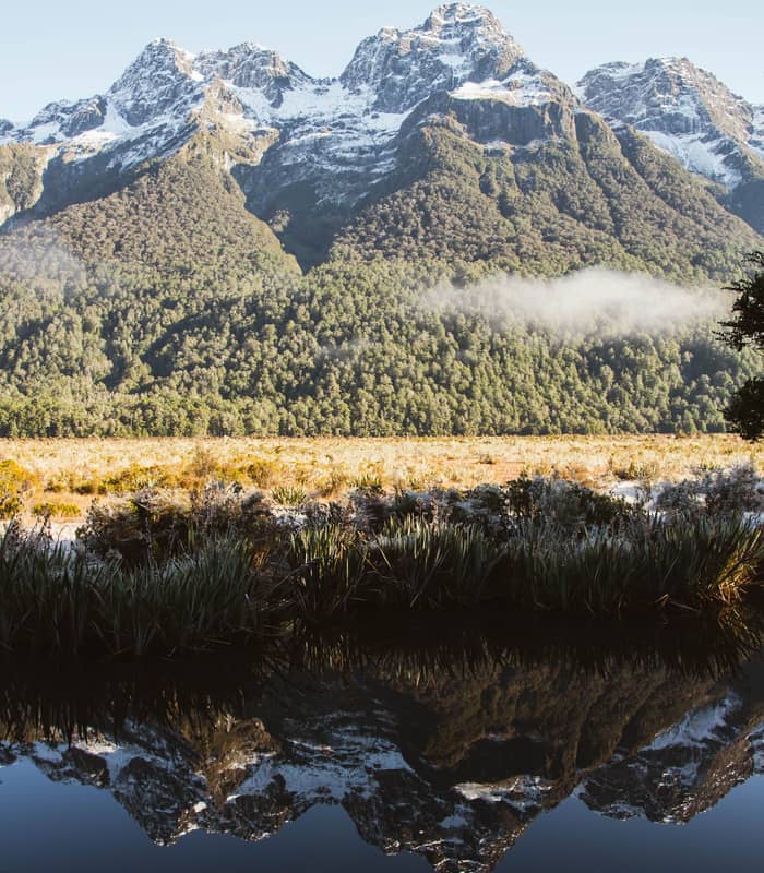Snowy mountains reflected in a still lake with forest and grassy vegetation around.