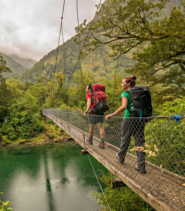 Two hikers with backpacks crossing a suspension bridge over a river in a forested mountain area.