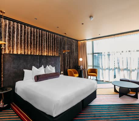 A wide view of a vibrant Habitat Twin room at Naumi Hotel Wellington with two beds, a pink sequined wall, a red velvet sofa, and a bold diagonally-striped carpet.