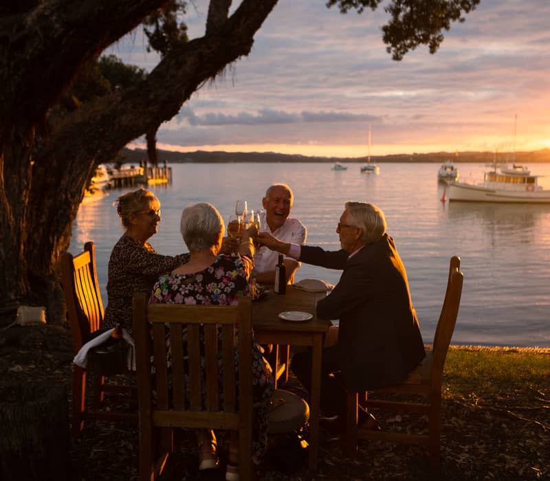 Four people toasting drinks at a table near a bay during sunset with boats on the water.