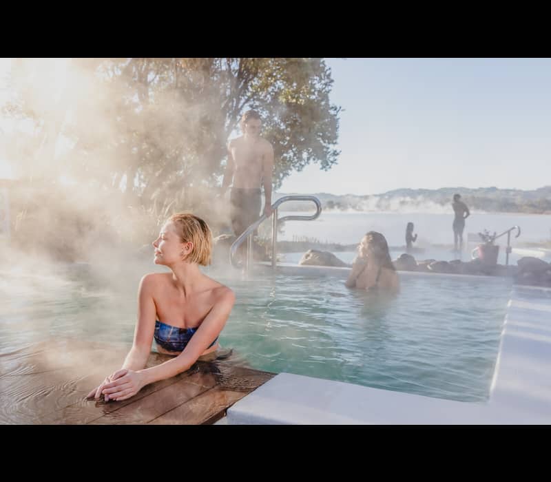 A woman relaxes in a steaming outdoor thermal mineral pool at sunset, with other bathers visible in the background against the misty shoreline of Lake Rotorua at the Polynesian Spa.