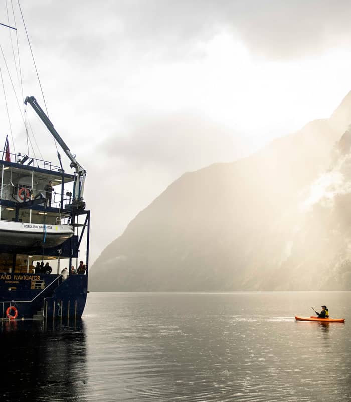 Ship Fiordland Navigator anchored in foggy Doubtful Sound with kayaker on calm water near mountains.