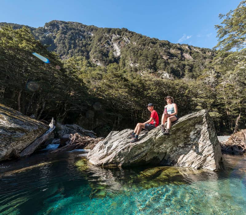Two people sitting on rocks by a clear blue river with forest and mountains in background.