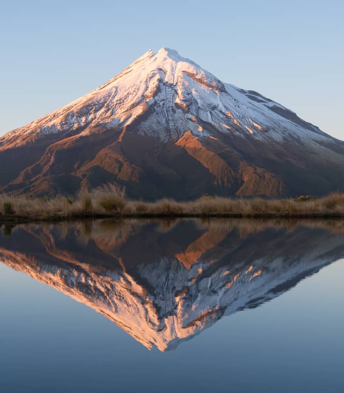 Snowcapped Mt Taranaki reflected clearly in a calm lake at sunset.
