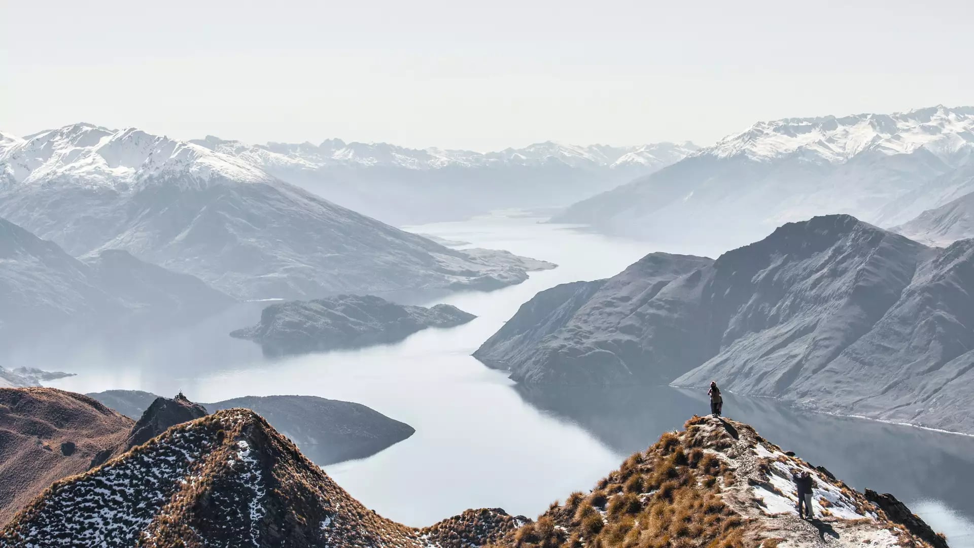 A person stands on a steep mountain peak overlooking a vast, winding lake surrounded by dramatic snow-capped mountain ranges.