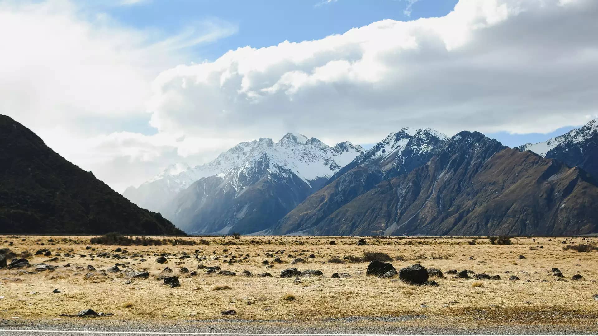 A wide roadside view looking across a rocky, golden tussock valley floor toward towering, snow-capped peaks of the Southern Alps under a partly cloudy sky.