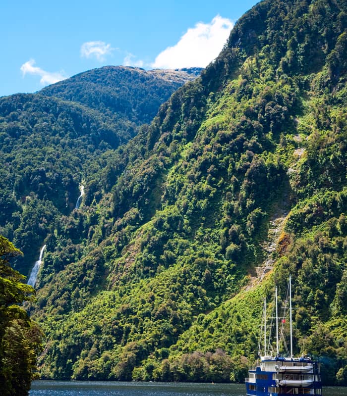 Blue cruise boat on water near green hills and waterfalls under blue sky
