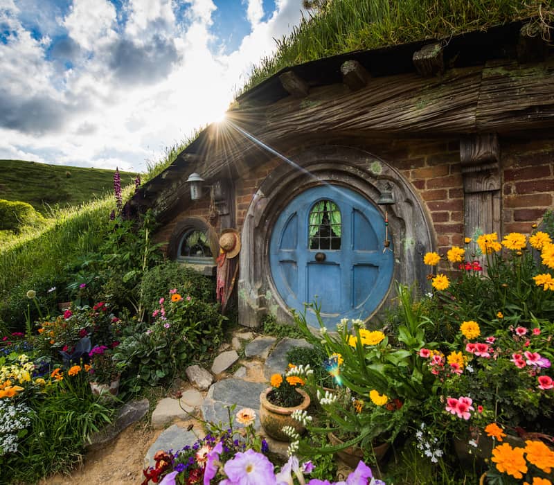 Blue round door of a hobbit house surrounded by colorful flowers and green grass with sun shining in the background.