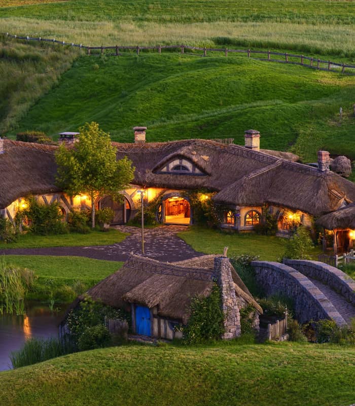 Thatched roof hobbiton cottage and stone bridge surrounded by green hills at sunset near a pond