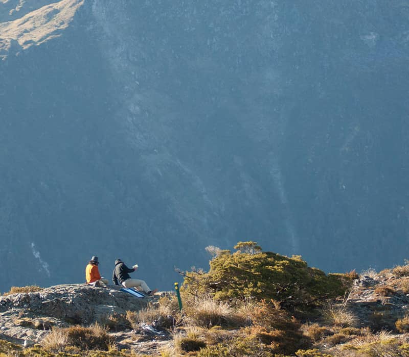 Two people sitting on a rocky mountain summit with scenic mountains in the background.