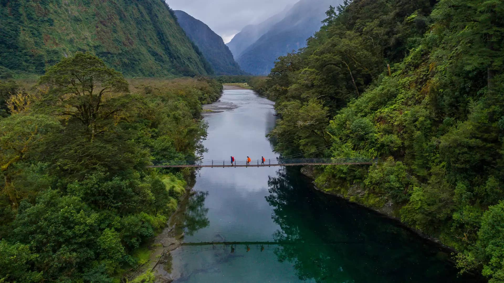 Three hikers crossing a suspension bridge over a river in a lush forested mountain valley.
