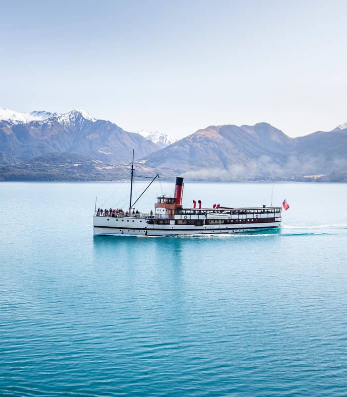 Steamboat cruising on blue lake water with snowy mountains in the background on a clear day.
