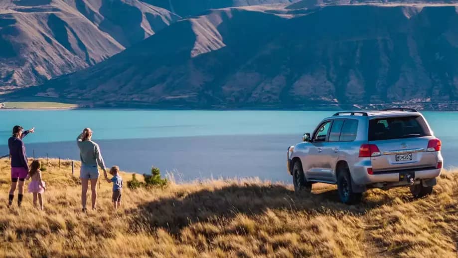 Family of four looking at lake and mountains with a parked SUV on grassy hill.