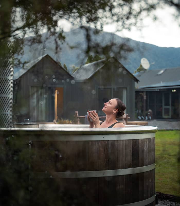Woman relaxing in outdoor wooden hot tub holding a cup with lodge and mountains in the background