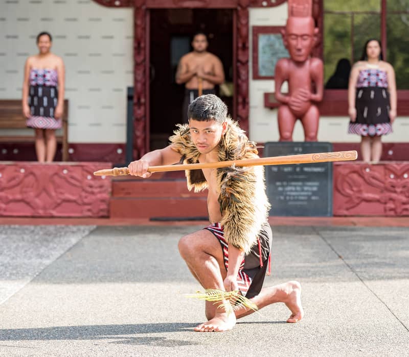 Maori man in traditional dress performing with wooden staff in front of carved statue and group of performers.