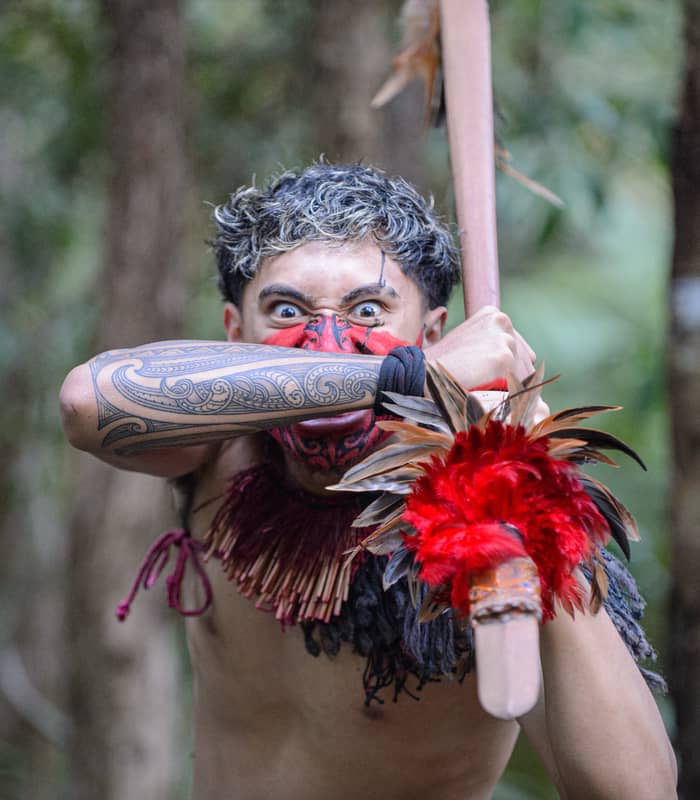 Māori warrior with tattoos and face paint holding a feathered weapon in a forest setting.