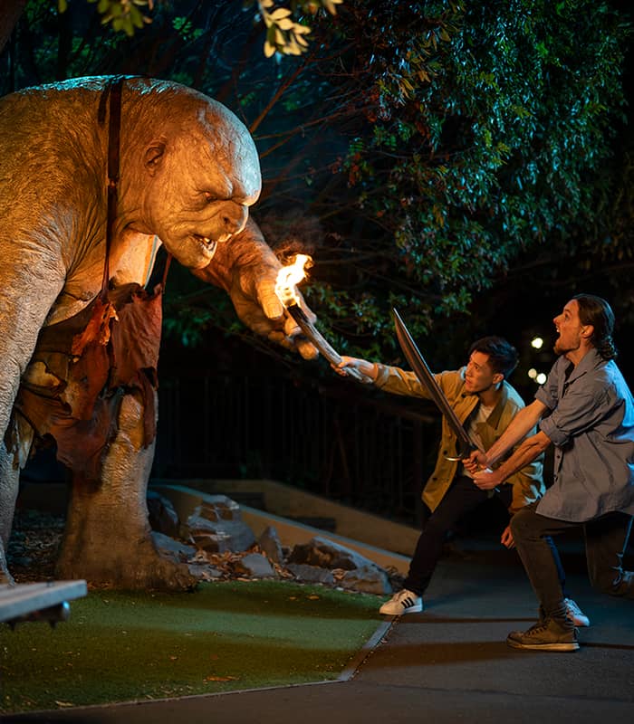 Two men with swords face a giant troll statue holding a flaming torch at night outdoors.